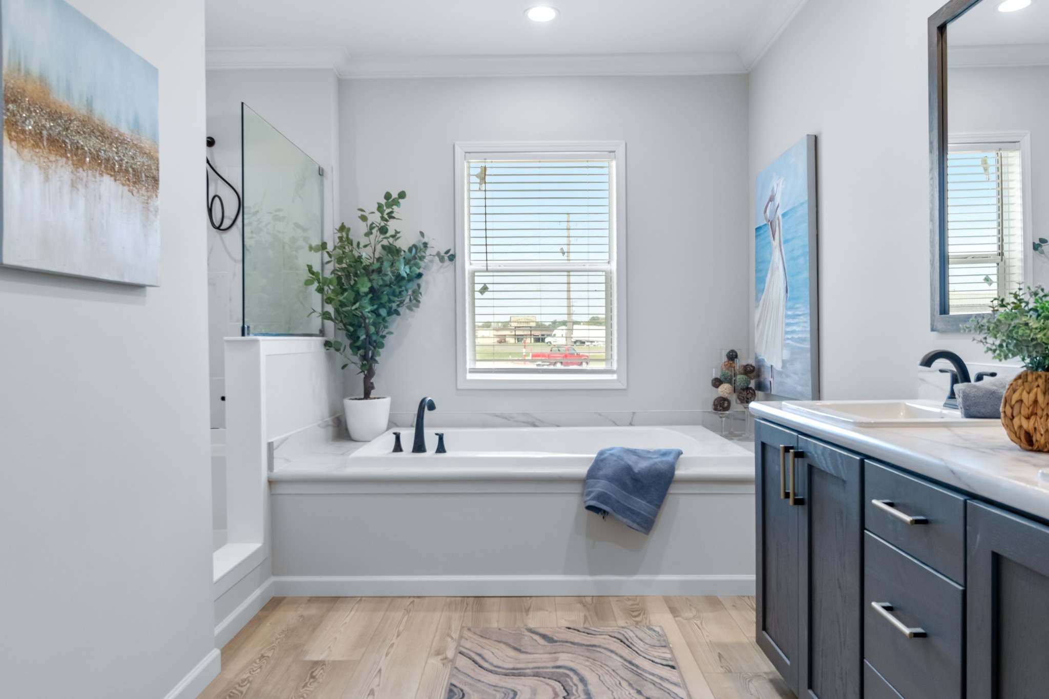 Modern bathroom with a white bathtub, blue towel, and a large window with blinds. Light wooden floor, dark cabinets, and potted plants add a serene touch.