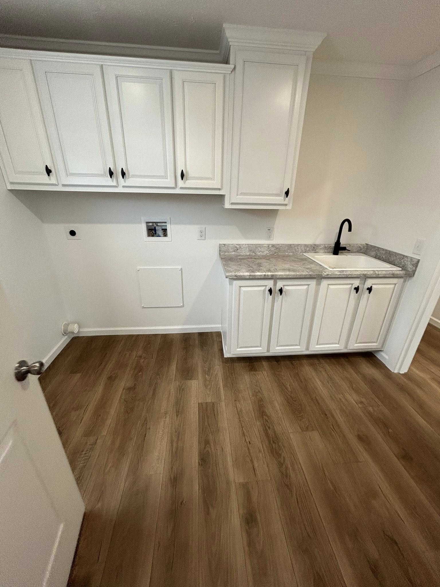 A small laundry room with white cabinets, a granite countertop, a black faucet, and a sink. The wood floor adds warmth to the clean, functional space.
