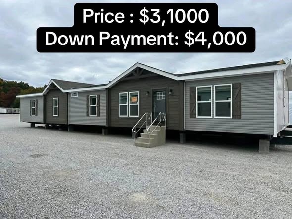 A gray mobile home with white trim sits on a gravel lot. Text overlay indicates a price of $31,000 and a down payment of $4,000. Overcast sky above.