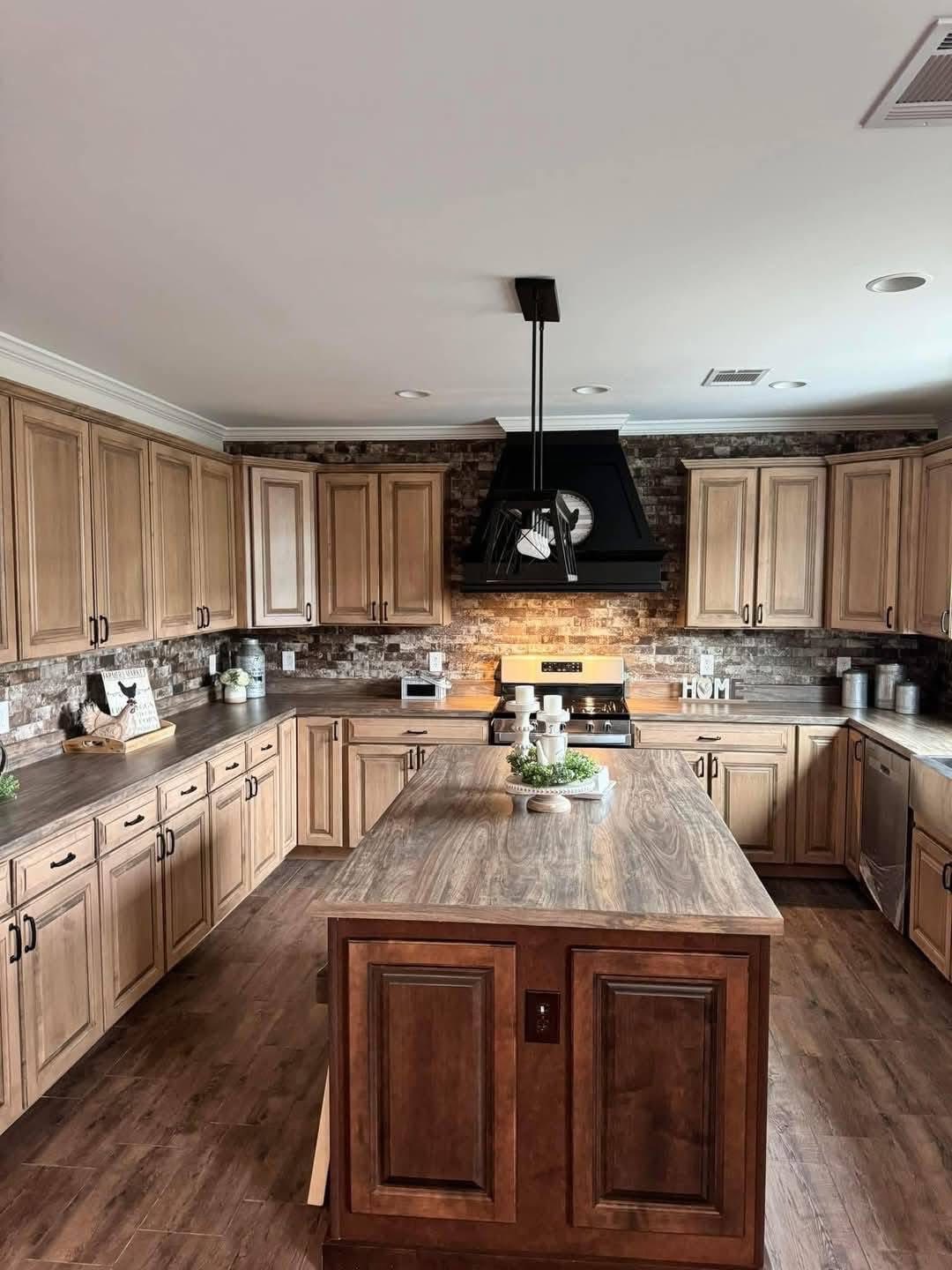 Alt text: Spacious kitchen with warm wooden cabinets, island, and dark wood floor. Brick backsplash adds rustic charm. Modern stove and hood in center.
