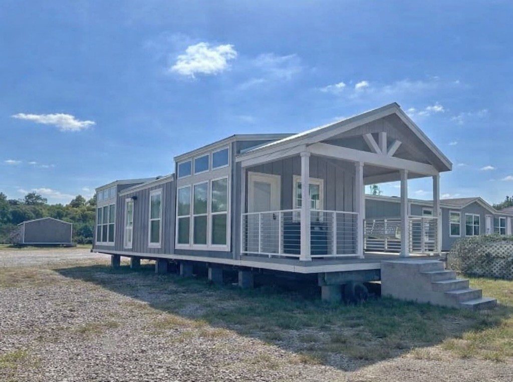 A modern, raised tiny house with a gray exterior, numerous large windows, and a small porch sits under a clear blue sky, conveying a sense of openness.