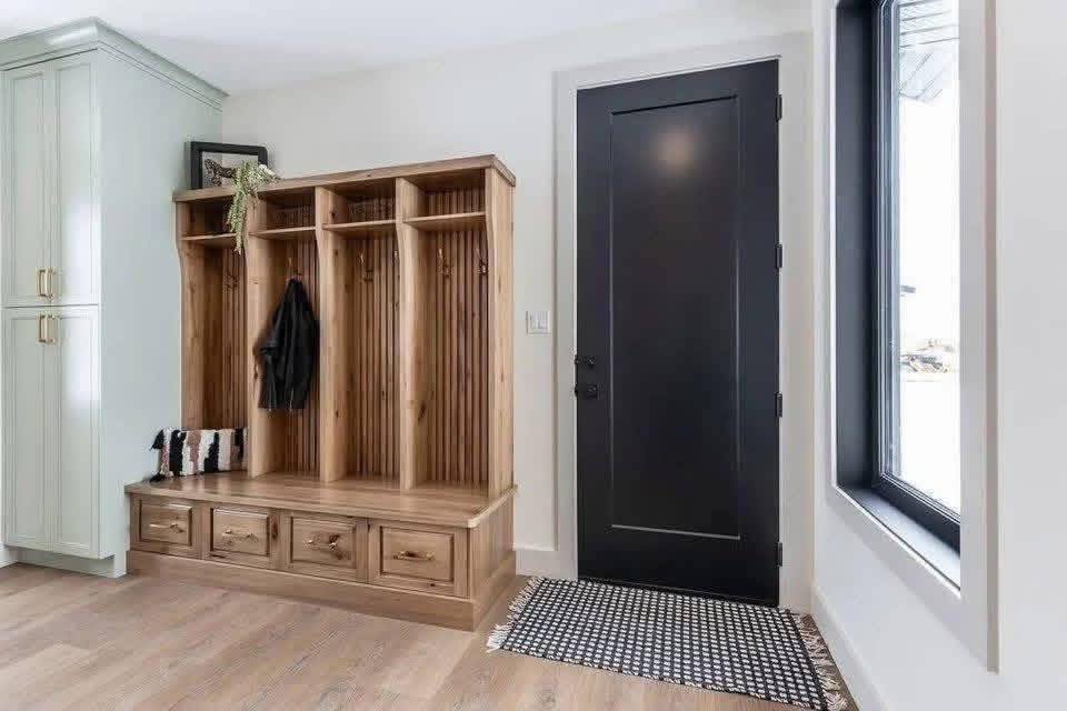 A tidy mudroom features a wooden bench with coat hooks and storage cubbies. A black door and a patterned rug add contrast to the bright, airy space.
