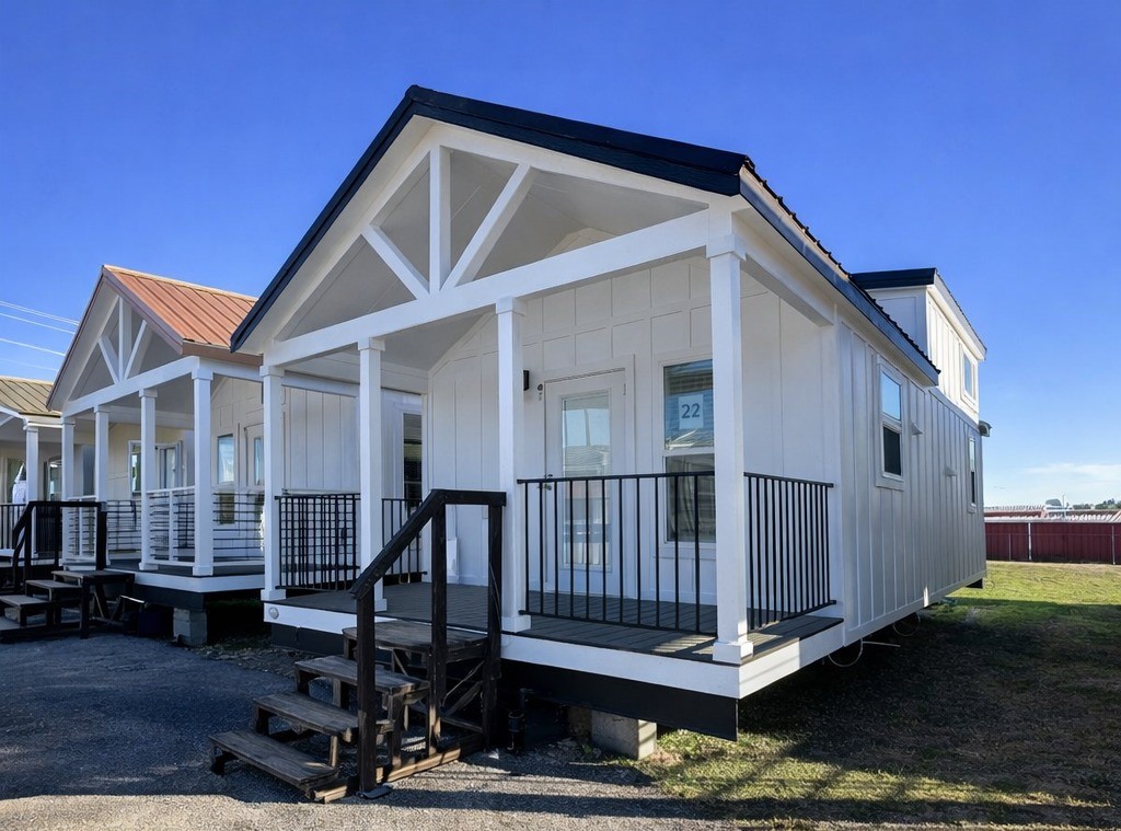A white tiny house with a black roof sits under a clear blue sky. It features a covered porch with railings and steps, exuding a modern, cozy vibe.