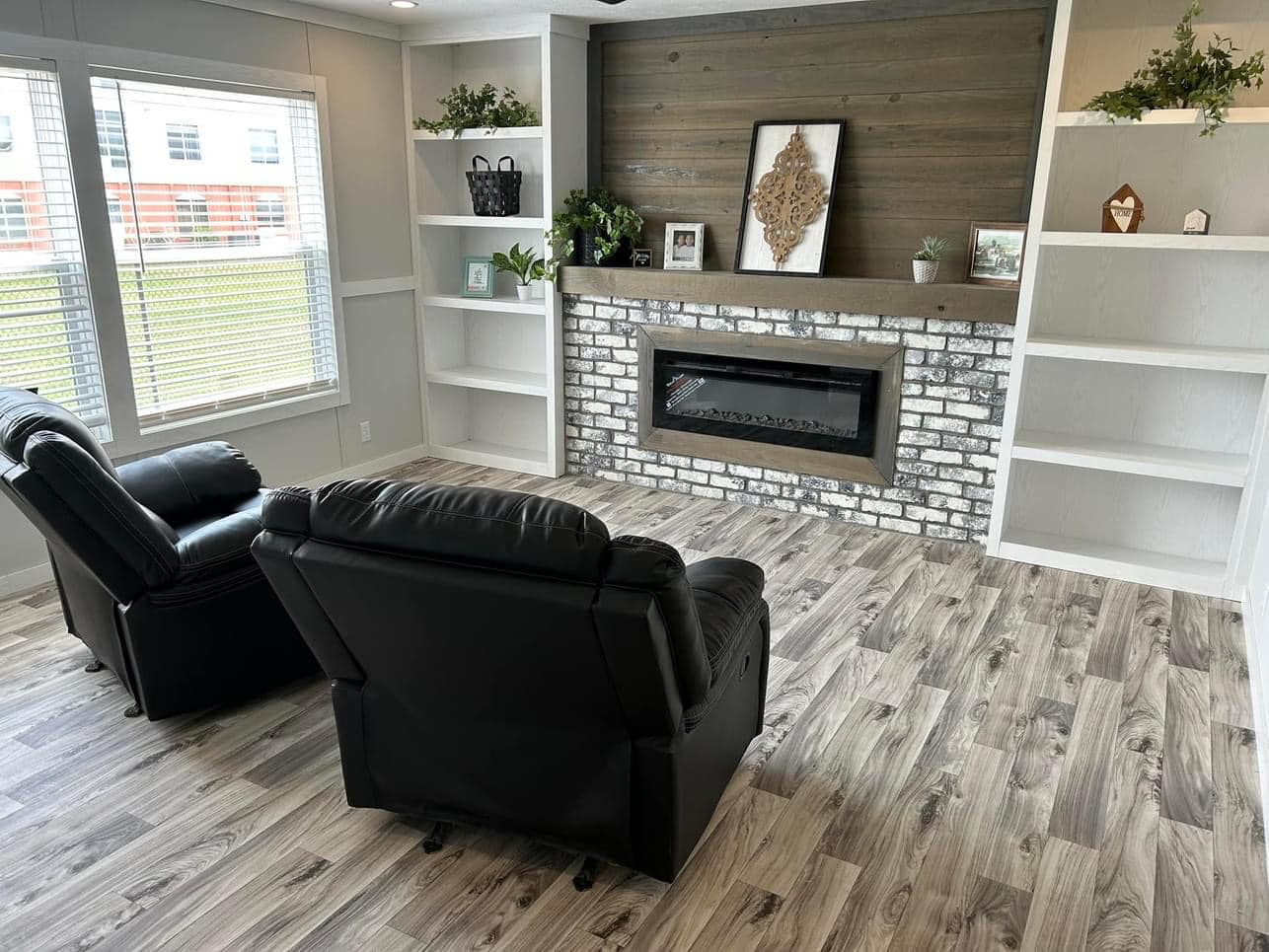 Cozy living room with two black recliners facing a fireplace set in a white brick wall. Wooden shelves hold plants and decor. Large windows add brightness.