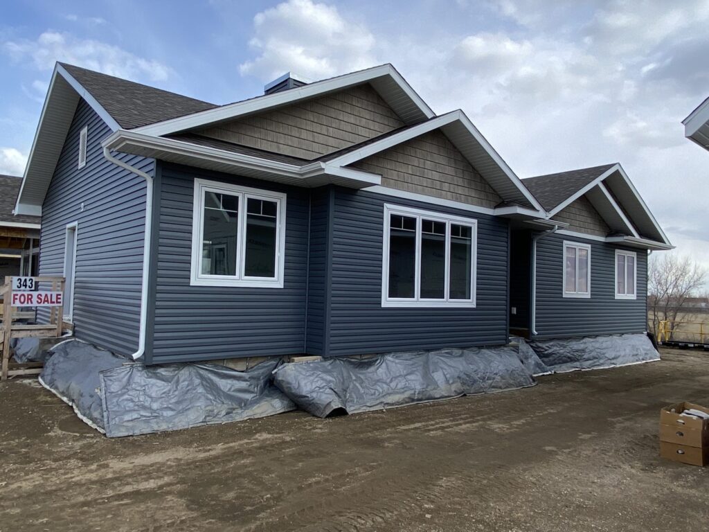 A newly constructed blue house with white trim and a gabled roof. "For Sale" sign on the left. Overcast sky. Calm and inviting atmosphere.