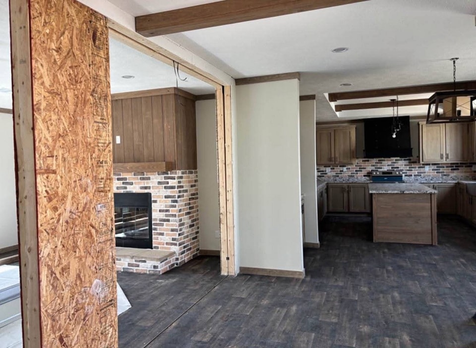 Spacious room with an unfinished doorway, featuring wood floors and a brick fireplace. Adjacent kitchen has wooden cabinets and dark countertops, exuding a rustic charm.