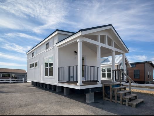 A small white house with a gabled roof and a front porch, elevated on a platform. Steps lead up to the porch, set against a clear blue sky.