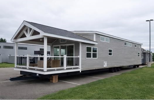 A modern mobile home with gray siding is parked on a paved area. It features a covered porch with white railings and large windows, under a cloudy sky.