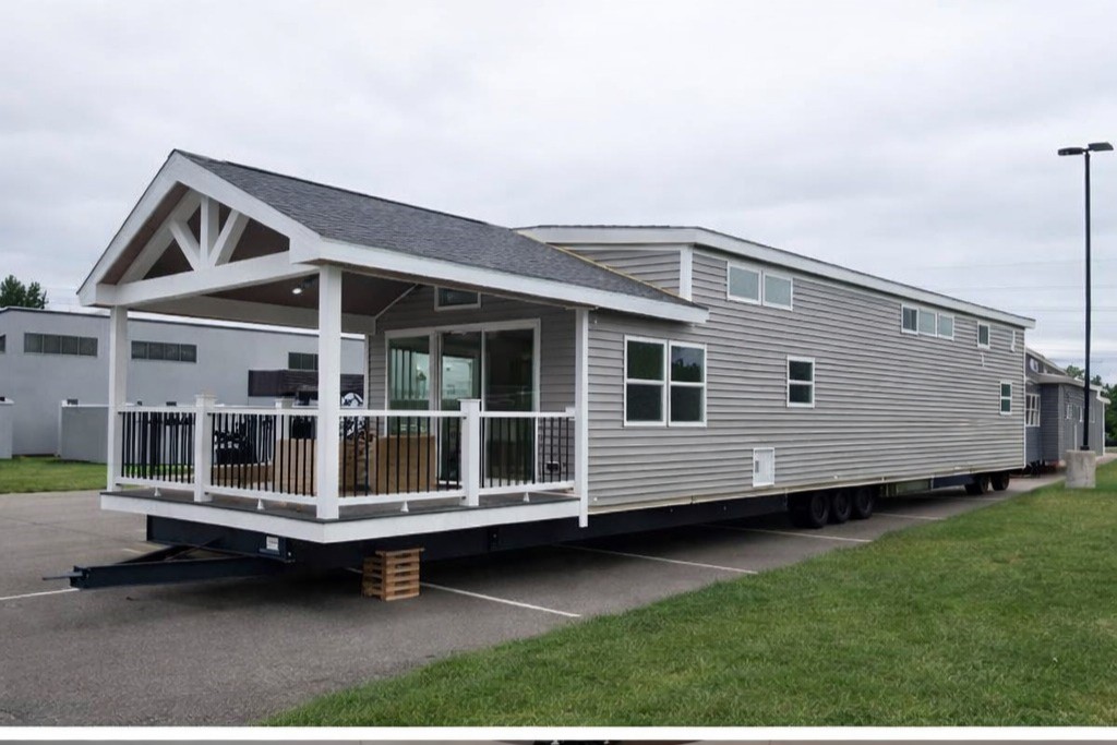 A long, modern mobile home with a covered porch is parked in a lot. It has gray siding, multiple windows, and sits on wheels, suggesting mobility.