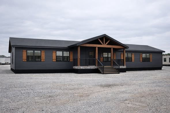 A modern, spacious, gray manufactured home with wooden accents stands on a gravel lot under a cloudy sky, evoking a sense of rustic charm.