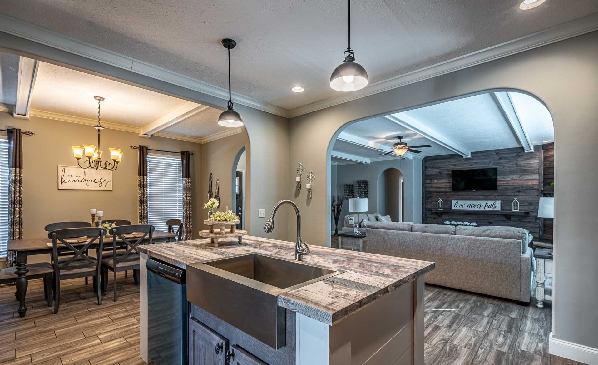 Modern open-concept kitchen and living room with rustic decor. A farmhouse sink island faces a living area with a large beige sofa, wood accents, and calming tones.