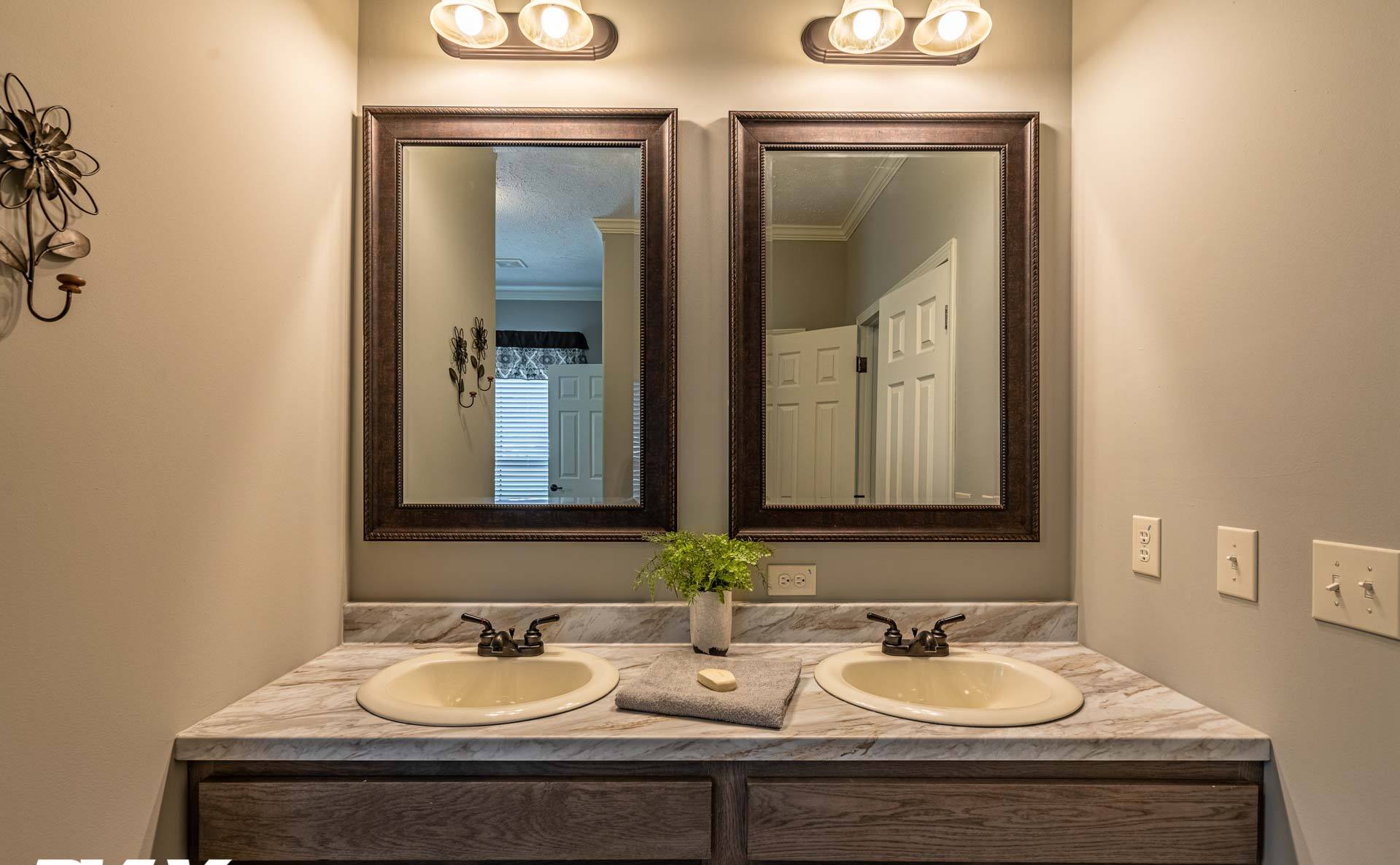Two sinks with marble countertops are topped by square-framed mirrors under warm lights. A small plant and towel add a modern touch to the bathroom.