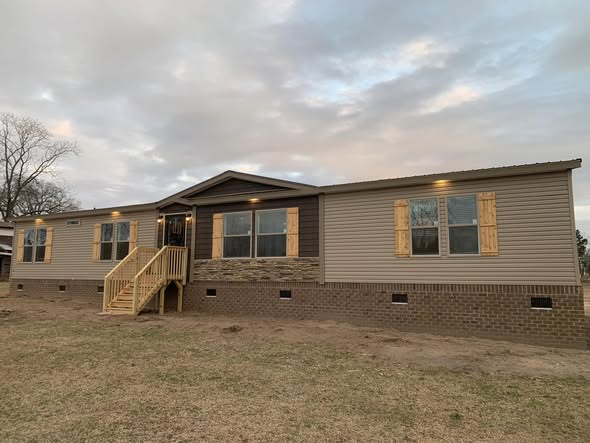 A large, single-story modular home with a beige exterior, brick foundation, and wooden shutters, set against a cloudy sky, creating a peaceful ambiance.