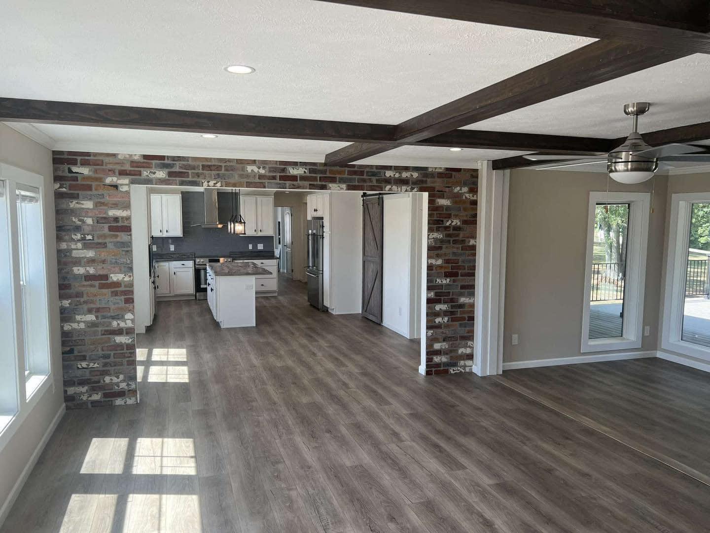 Spacious room with exposed brick wall and wooden beams, featuring a kitchen with white cabinets and a center island. Large windows offer natural light.