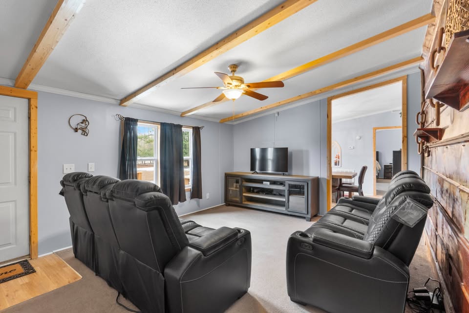 Cozy living room with black leather recliners facing a TV on a wooden stand. Ceiling fan above and a large window with curtains to the left. A dining area is visible through an open door. Warm, inviting atmosphere.