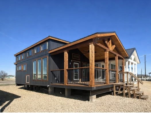 Modern tiny house with dark panels and a wooden porch under a clear blue sky. Features large windows, wood accents, and gravel surroundings, evoking a cozy, minimalist feel.