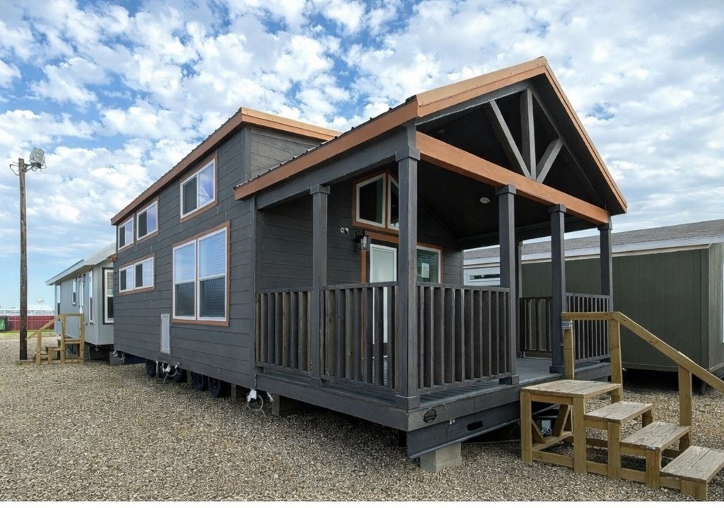 A tiny house with dark wood siding and a small porch sits on gravel under a partly cloudy sky. Its elevated design includes wooden steps.