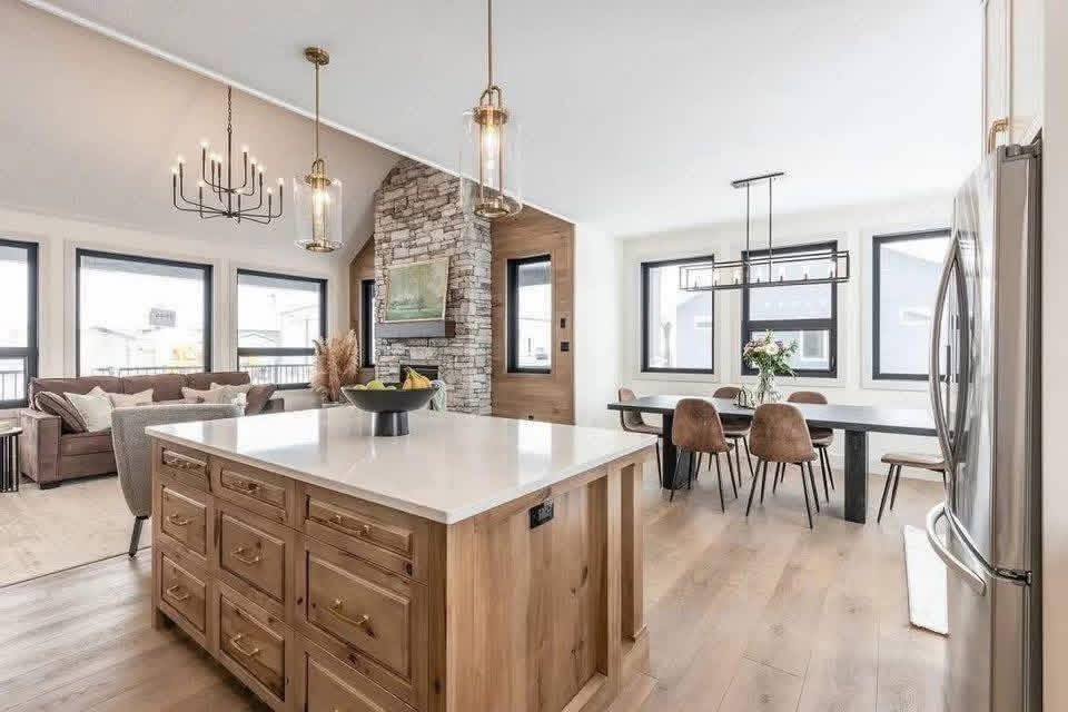 Modern open-concept living space with a large kitchen island and wooden cabinets. Features a dining area, cozy living room, and stone fireplace. Bright and airy.