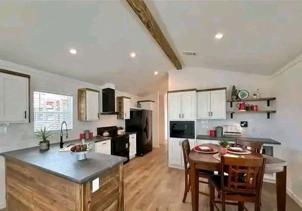 Warm kitchen with wooden floors, modern white cabinets, and black appliances. A wooden dining table set for four adds a cozy, welcoming feel.