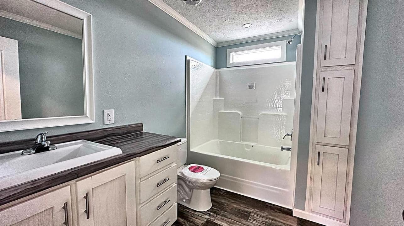 A modern bathroom with light blue walls, featuring a dark wood vanity, white sink, bathtub with shower, and tall white cabinets on a wooden floor. Bright and clean ambiance.