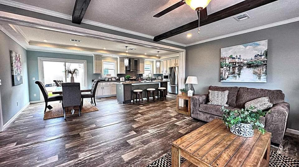 Open-concept living space with hardwood floors. Cozy brown sofa, wooden coffee table, modern kitchen with bar stools, dining area, and soft lighting.