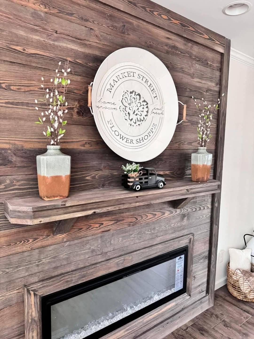 Wooden wall with a "Market Street Flower Shoppe" sign, flanked by two vases with white flowers. A small truck decor piece sits on a shelf above an electric fireplace.