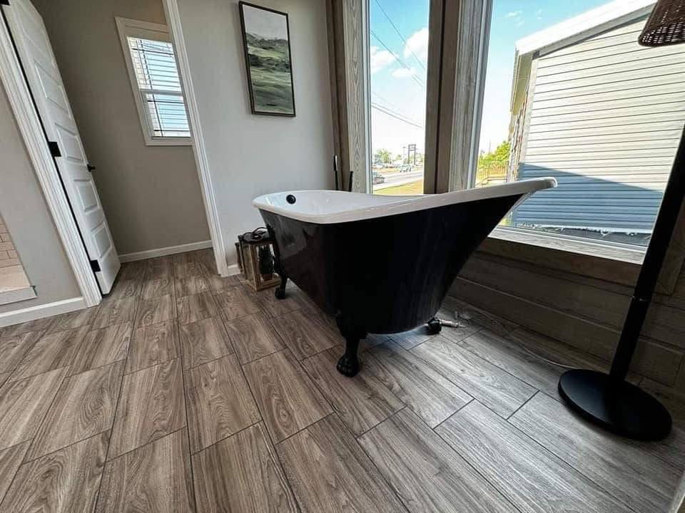 A black freestanding clawfoot bathtub sits near large windows showcasing an outdoor view. The room has wood-patterned tile flooring and neutral walls, creating a serene and modern atmosphere.
