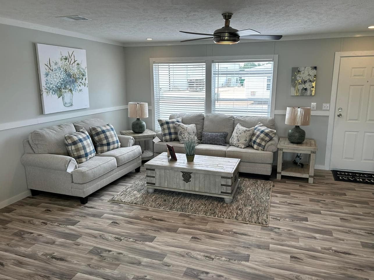 Cozy living room with neutral tones, featuring two sofas with plaid cushions, a rustic wooden coffee table, soft lighting from two lamps, and floral wall art.