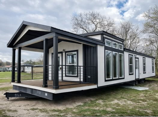 Modern mobile home with black and white exterior, large windows, and a covered porch set on a grassy area under a partly cloudy sky.