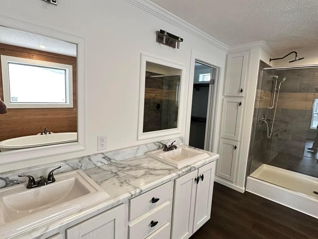 Modern bathroom features a marble countertop with dual sinks, white cabinets, a glass-enclosed shower, and wood-paneled wall with a bathtub.