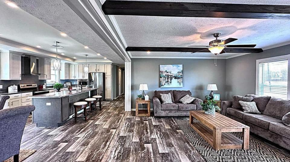 Modern living room with rustic wood floors, gray sofas, and a wooden coffee table. Open kitchen features bar stools and pendant lights. Cozy, inviting atmosphere.