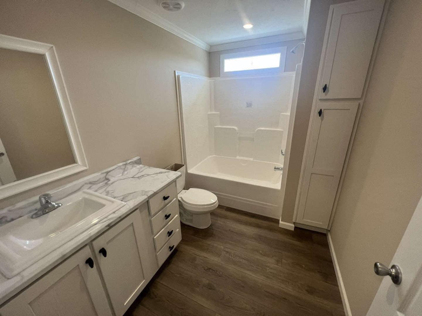 A modern bathroom with beige walls, featuring a white bathtub under a narrow window, a marble countertop with sink, wooden floor, and built-in cabinets.