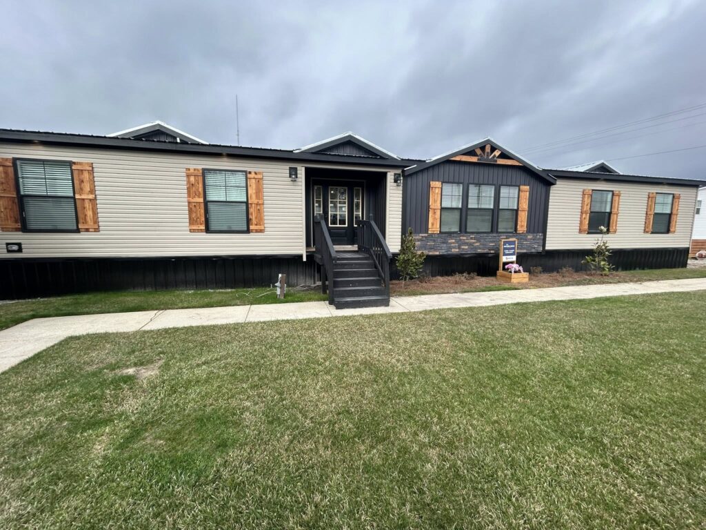 Modern modular home with light siding and wooden shutters. A black front porch and stone accents stand out against a cloudy sky and grassy lawn.