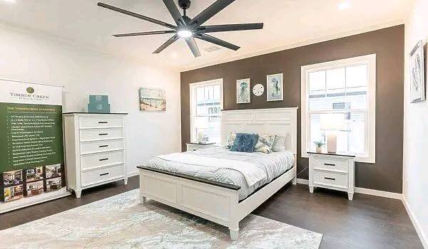 Modern bedroom with white furniture, including a bed, dresser, and nightstand. Brown accent wall, large ceiling fan, and artwork create a serene ambiance.