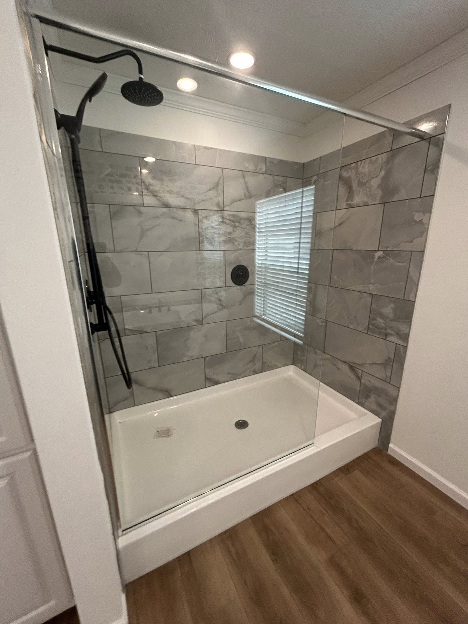 Modern shower with gray marble tiles, glass door, and black fixtures. A small window with blinds brings natural light, while wood flooring adds warmth.
