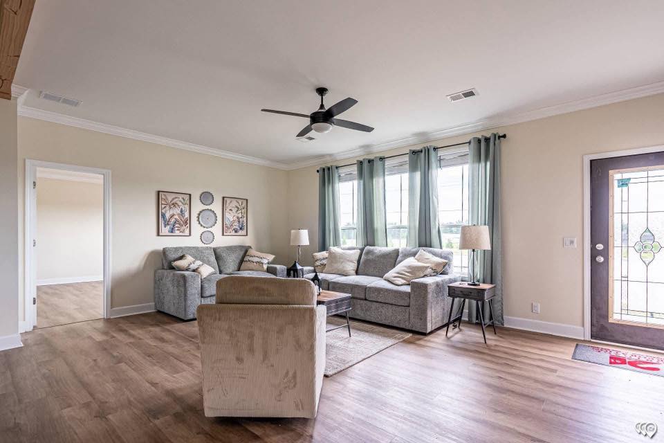 Spacious living room with gray sofas, floor lamps, and a ceiling fan. Large windows with teal curtains let in natural light. Wooden floor adds warmth.