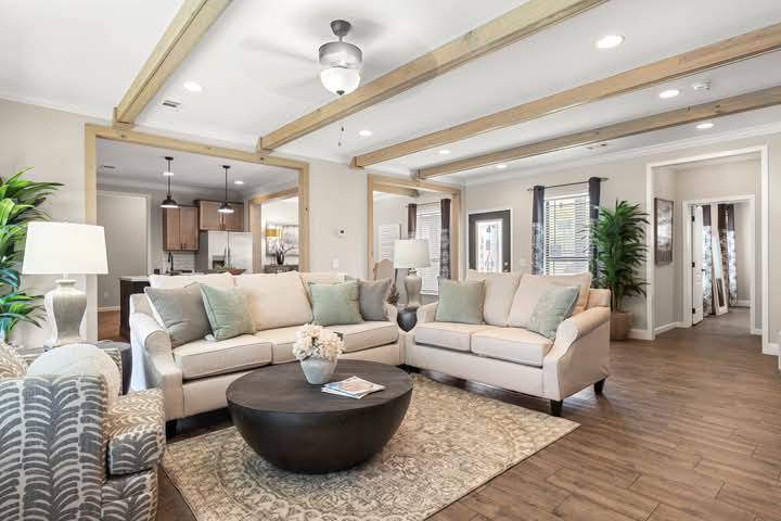 Spacious living room with beige sofas adorned with green cushions, a round black coffee table, wooden beams on the ceiling, and warm lighting. Cozy and inviting.