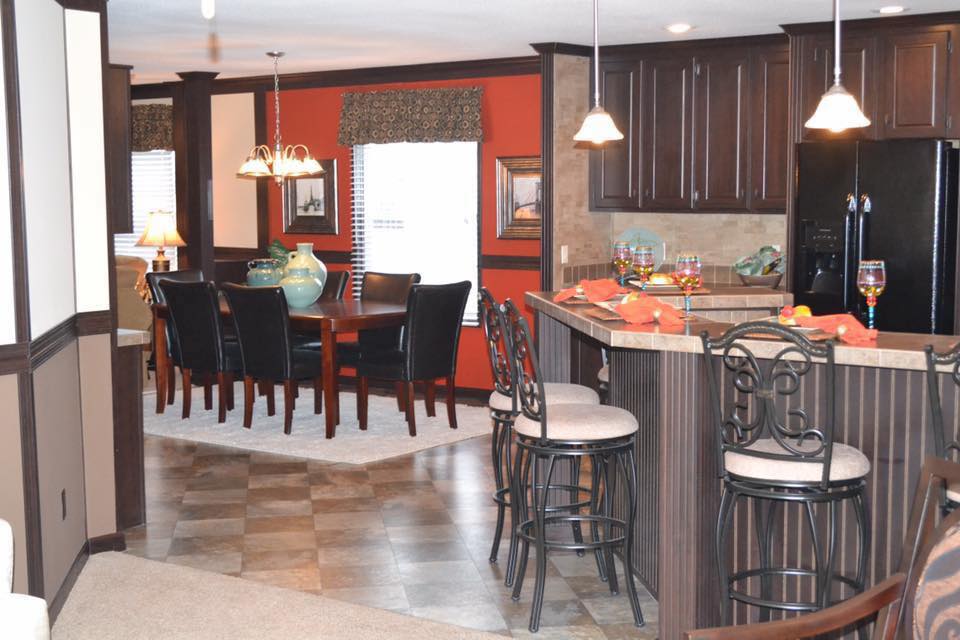Warm, inviting kitchen and dining area with dark wood cabinets, red accents, and elegant lighting. Bar stools and a dining table create a cozy atmosphere.