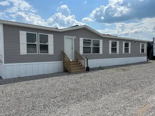 A gray mobile home with white trim and shutters sits on a gravel lot under a partly cloudy sky. Wooden steps lead to the front door, creating a peaceful scene.
