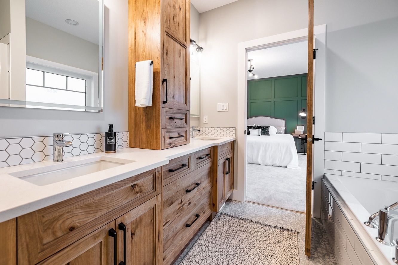 Modern bathroom with wooden cabinets, white countertops, hexagonal tile backsplash, and a towel. The door opens to a bedroom with a green accent wall.