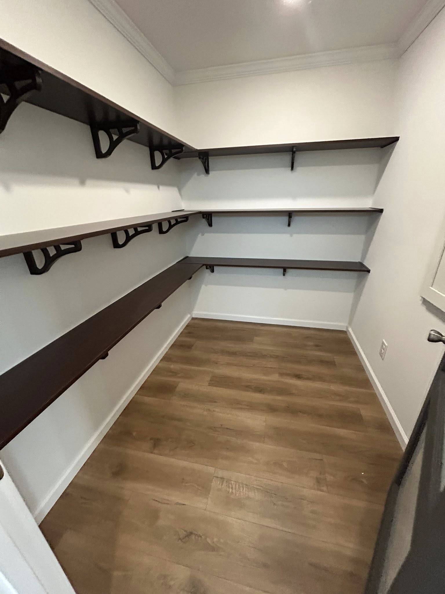 Empty walk-in pantry with dark wooden shelves on three white walls, creating a clean and organized look. The floor is light wood, adding warmth.