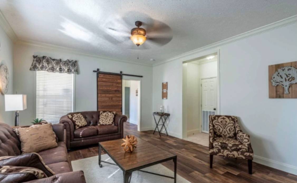 Cozy living room with brown sofas, patterned armchair, and a wooden table. Warm lighting from ceiling fan, rustic barn door, tree wall art, and wood flooring.