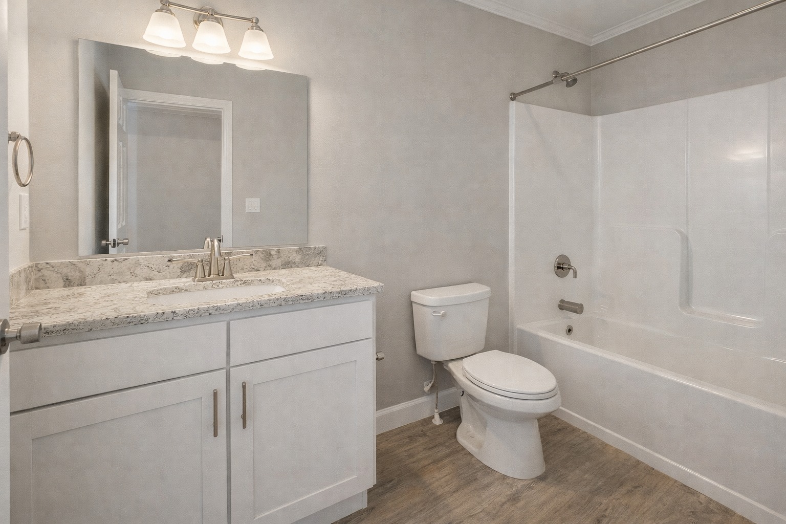 Modern bathroom with white vanity and granite countertop, large mirror, and three light fixtures. Bathtub and toilet with light wood flooring. Simple and clean.