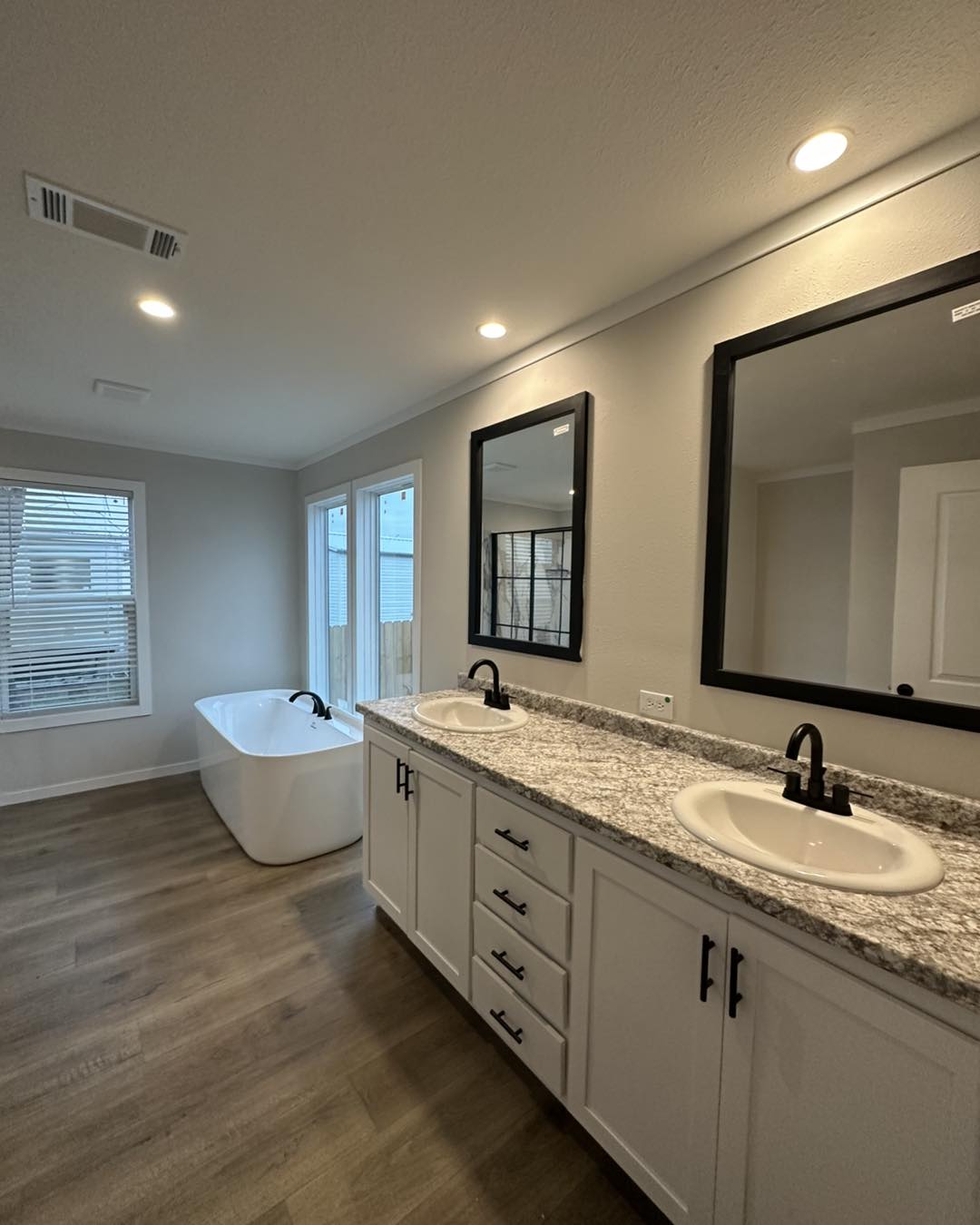 Spacious empty room with light gray walls and brown wooden floor. Ceiling fan with light hangs overhead. Two windows with blinds bring in soft light.