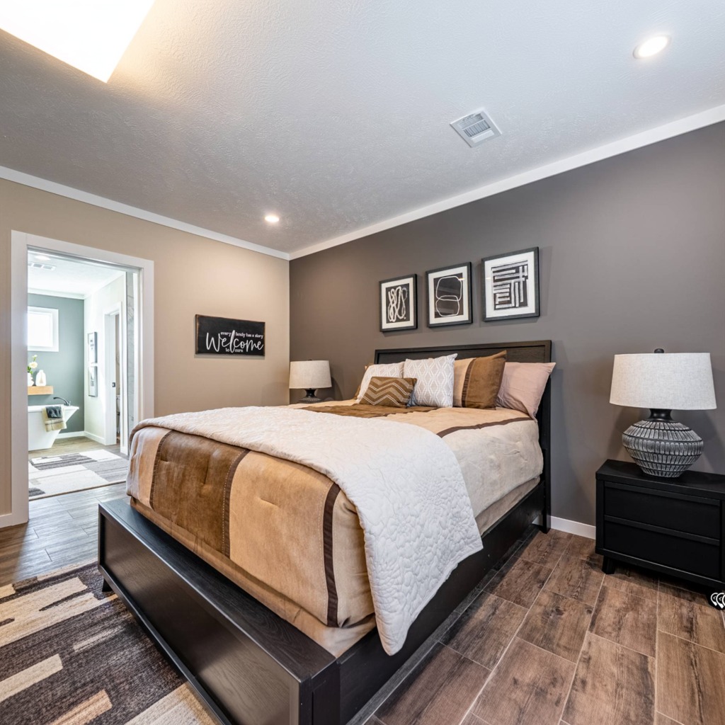 Modern bedroom with a large bed featuring beige and white bedding, flanked by two lamps on dark nightstands. Abstract art hangs on a gray wall. A doorway leads to a lit bathroom.