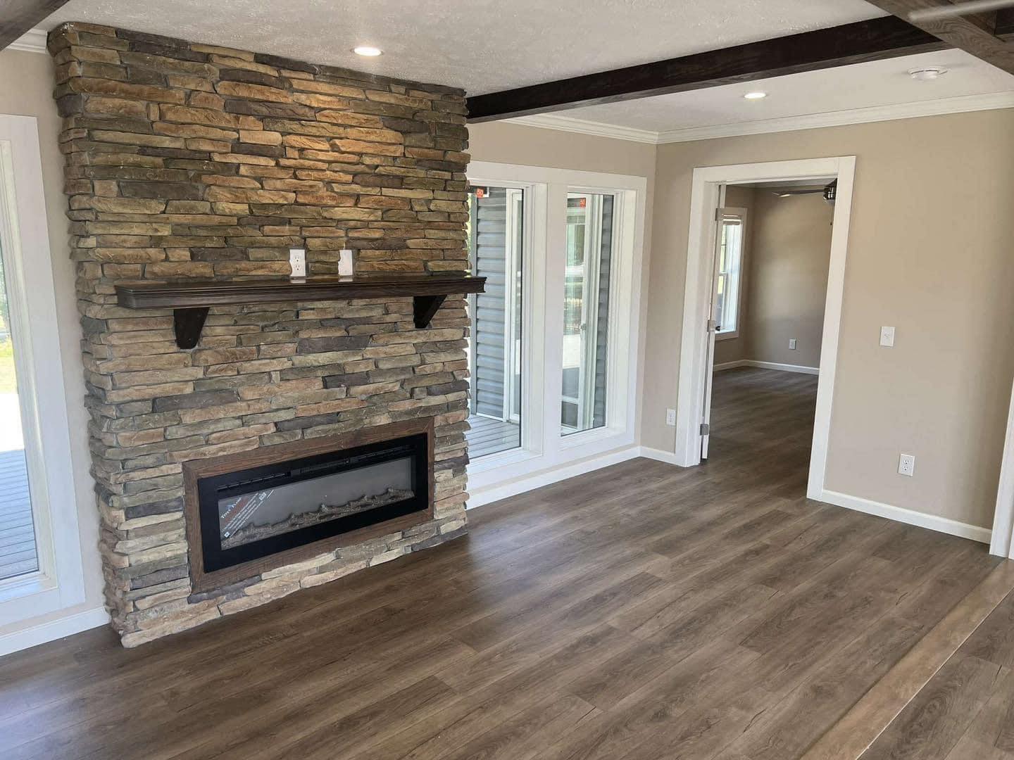 Cozy living room features a rustic stone fireplace with a wood mantel, light wood flooring, neutral walls, and bright windows, creating a warm ambiance.