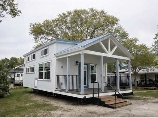 Modern tiny house with a blue door sits on a grassy lot. It features a porch with railings and large windows, surrounded by trees. Overcast sky above.