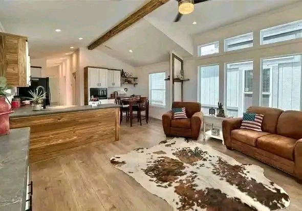 Spacious living room with wooden beams, tan leather sofas adorned with American flag cushions, cowhide rug, and large windows, conveying a cozy rustic feel.