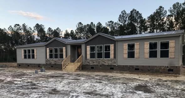 A beige modular home with stone accents and wooden shutters sits on a cleared plot, surrounded by tall trees, under a clear, evening sky.