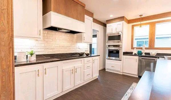 Modern kitchen with white cabinets, dark countertops, and brick backsplash. Stainless steel appliances include double ovens. Bright lighting and wood accents create a warm, inviting atmosphere.